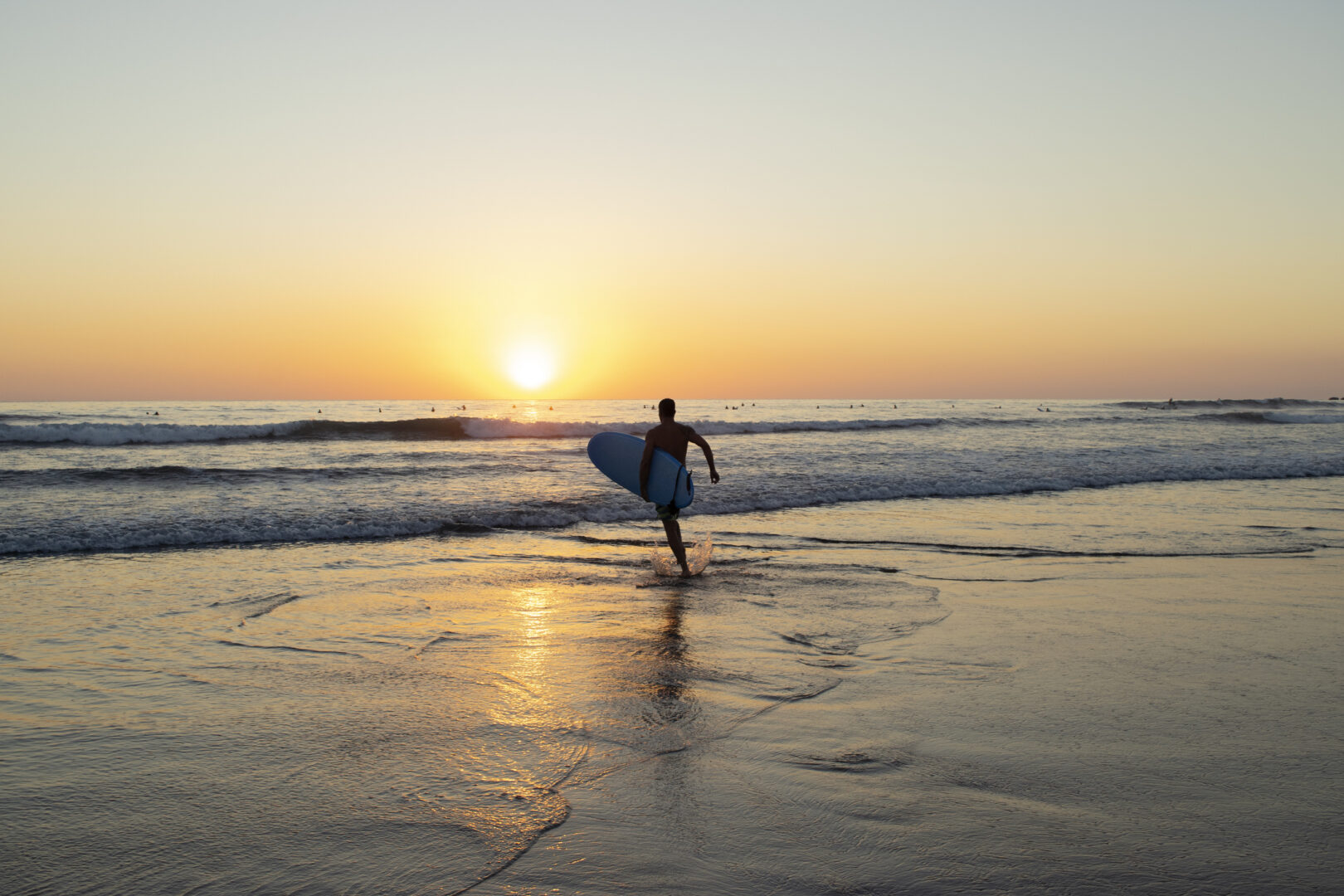 A male surfer with his surf board under his arm is running toward the waves to enter the water at sunset in Nosara, Costa Rica