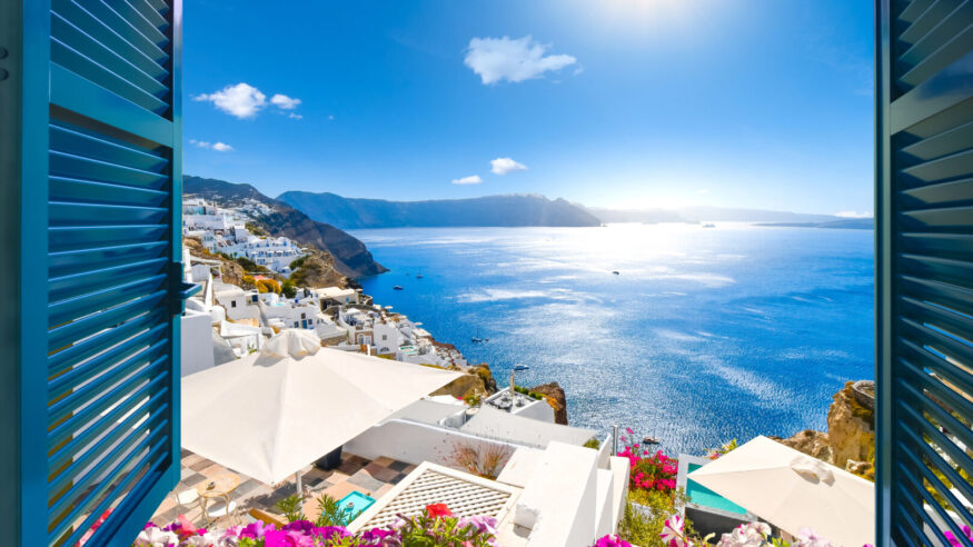 View from an open window with blue shutters of the Aegean sea, caldera, coastline and whitewashed town of Oia, Santorini, Greece.